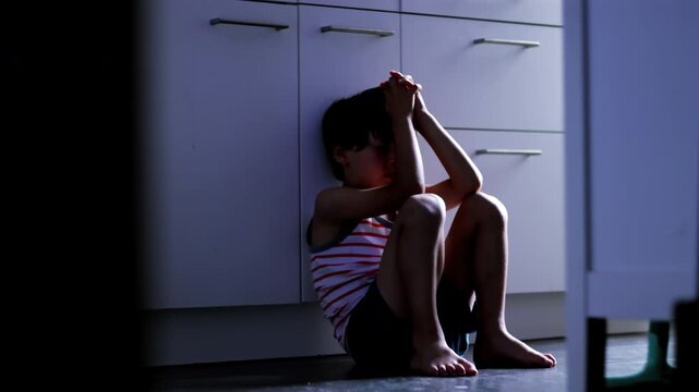 Child sitting on kitchen floor with hands clasped over head expressing overwhelming stress anxiety and childhood trauma in domestic environment suggesting fear and emotional breakdown