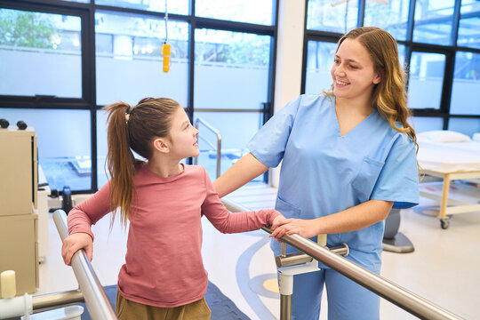 Pediatric physiotherapy session, therapist assisting child with rehabilitation exercises on parallel bars, motor skills training at pediatric rehab center, physical therapy session