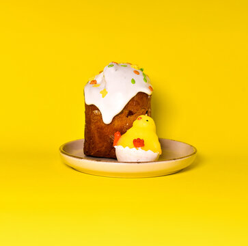 stive Easter scene with a glazed paska cake and a cute chick candle on a yellow plate. Vibrant monochromatic composition on a yellow background. Creative holiday food photography.