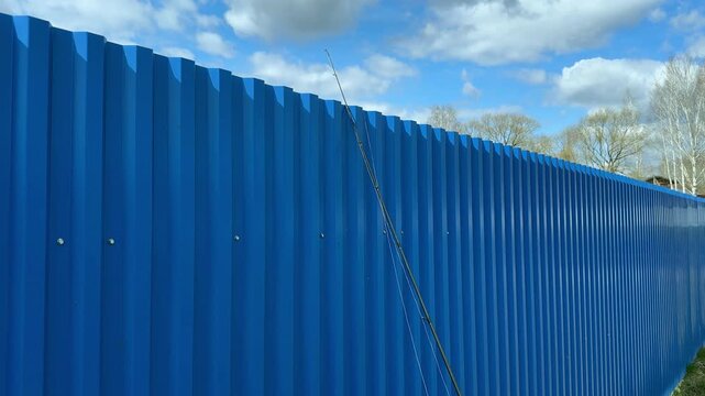 Blue corrugated fence lines a roadside under a bright sky. A long metal barrier stretches into the distance beside trees and clouds. Outdoor infrastructure and construction background with a