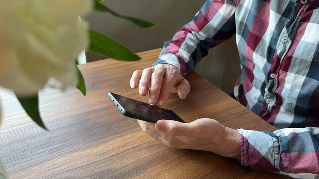 Senior man using a smartphone at a wooden table. Hands hold the mobile device during casual indoor browsing. Digital communication, online access, and home technology use..