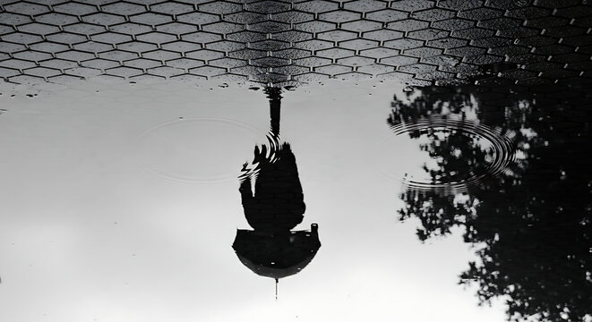A solitary figure holding an umbrella is reflected upside down in a puddle on a wet, tiled surface.