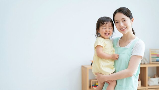 明るく清潔な室内で幼い子どもを抱いて微笑む女性保育士のポートレート。保育園、託児、子育て支援、幼児教育、ベビーシッター、育児サービス、福祉、家庭的なケア、安心感のある保育環境を表現した広告向け人物素材