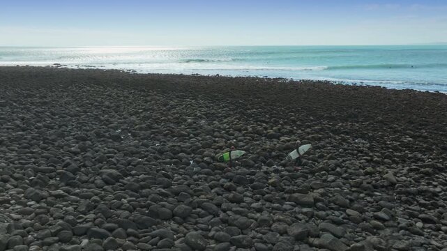 Surfers walk across a rocky shore towards the ocean on a sunny day in Raglan, Waikato, New Zealand. The surfers are preparing to catch waves.