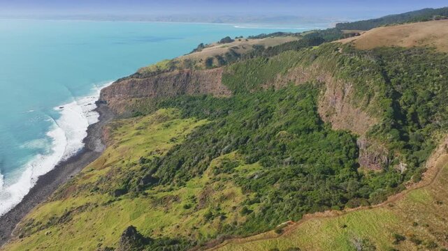 Rugged cliffs meet the turquoise ocean along the scenic coastline of the Raglan, Waikato, New Zealand. Lush green vegetation covers the rolling hills, showcasing the natural beauty.