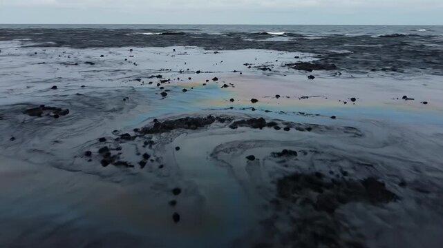 Thick black crude oil and iridescent rainbow fuel slick covering the ocean surface, showing an environmental disaster and severe marine water pollution under an overcast sky.