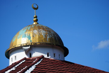 Ancient weathered gold mosque dome with patina, featuring a crescent moon and star finial. Located on a red tiled roof against a clear blue sky. Symbol of Islamic architecture and culture. © Nungkysoer