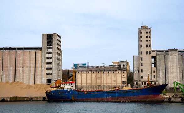 Cargo ship docked near old warehouses with sand piles on the shore in a busy port area
