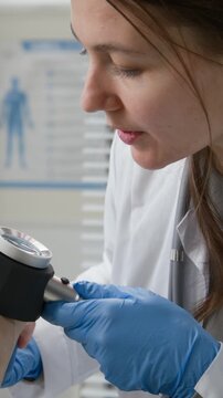 Vertical close up of young adult female dermatologist examining skin on forearm of elderly woman with dermascope during preventive screening in consultation room of clinic