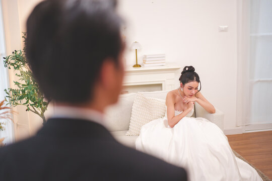 Sad Asian bride sitting on sofa with groom in foreground