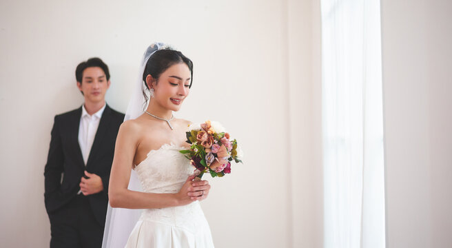 Portrait of Asian bride looking at bouquet with groom behind