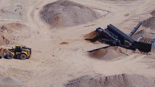 Aerial drone view shows a CAT wheel loader working among gravel and sand piles. Conveyors on a mobile plant form stockpiles as tyre tracks mark the arid ground.
