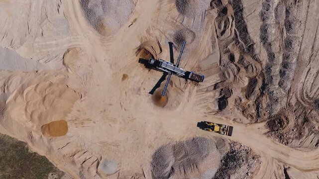 Aerial view of an open pit aggregate quarry with a cross shaped conveyor, cone stockpiles, a yellow loader on a dirt road, and crushed rock discharging in daylight.