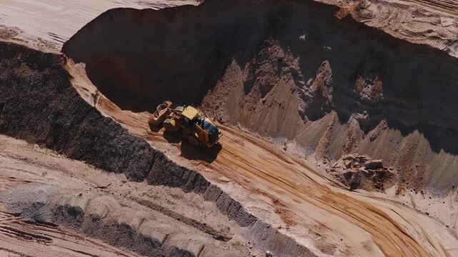 Aerial view shows a yellow wheel loader grading a haul road in a steep open pit quarry, with layered cut faces, berms, parallel tire tracks, and spoil piles.