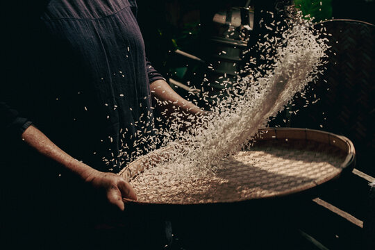 Action shot of a person traditionally winnowing raw rice grains using a bamboo tray, separating husks with motion blur in a dark rustic kitchen setting, symbolizing organic harvest and farming.
