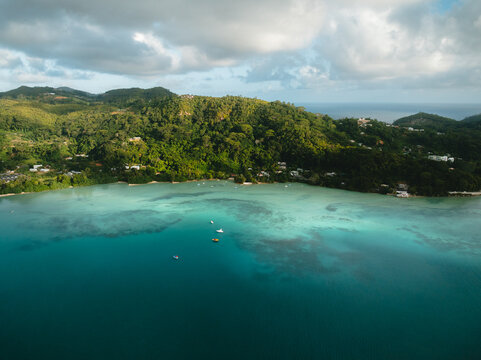 Aerial view of Anse A La Mouche with turquoise waters and lush green hills under a cloudy sky in Seychelles.