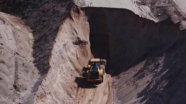 Aerial view shows a yellow wheel loader moving in a narrow trench, leaving fresh tread marks and a deep shadow, steep layered bench walls, and haul road tire tracks.