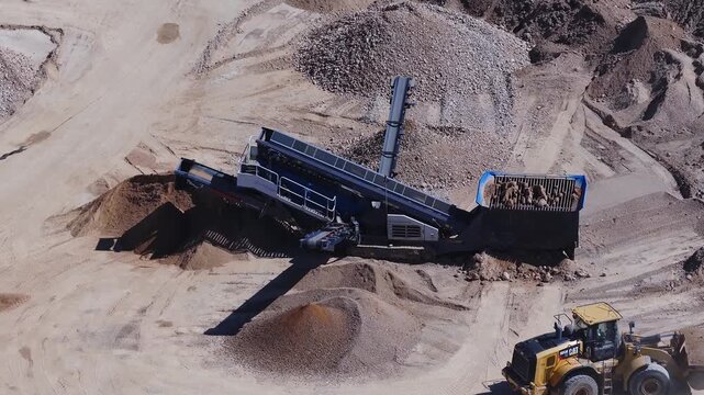 Aerial midday view of a quarry as a loader feeds a blue topped hopper and a conveyor places crushed rock into conical stockpiles, with deep shadows and clear tire tracks.