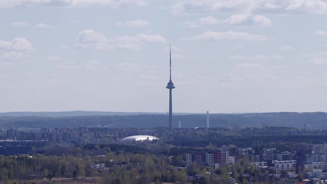 Aerial pan over Vilnius, Lithuania centers Vilnius TV Tower above low apartment blocks, silver domed arena, spring trees, soft midday light, telephoto flattening.