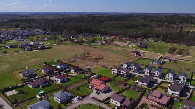 High wide aerial shot over Vilnius suburbs shows modern houses, solar panels, pools, greenhouses, allotments, active builds, and a forest belt on a clear spring day.