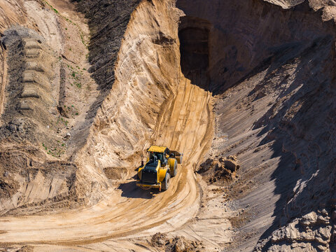 Aerial view shows a yellow wheel loader on a narrow winding haul road in an open pit mine, fresh tire tracks, layered sediment, and a shadowed adit under midday light.