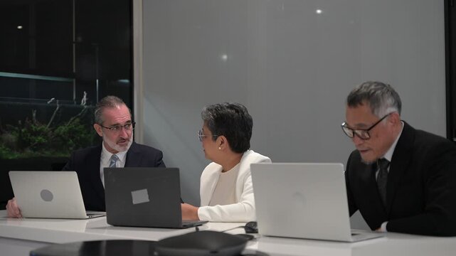 Diverse senior business executives discussing global corporate strategy during a late night board meeting in a modern office. Professional management team collaborating and negotiating with laptops.