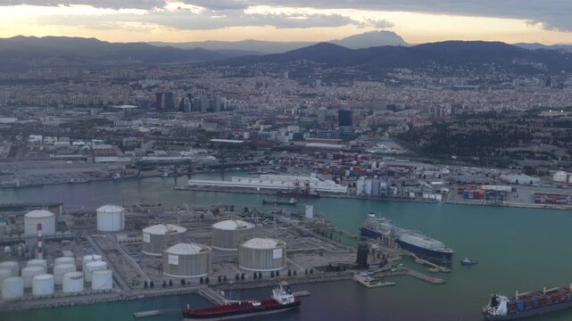 Aerial view of barcelona's industrial port at dawn, showing lng storage tanks, refineries, and container terminals with cargo ships and the city skyline in the background