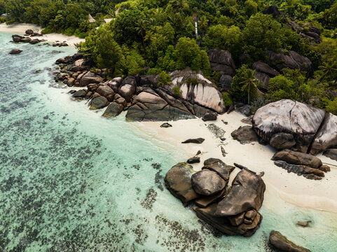 Aerial view of the granite rock formations and white sand beach with turquoise water and lush tropical forest in Takamaka, Seychelles.