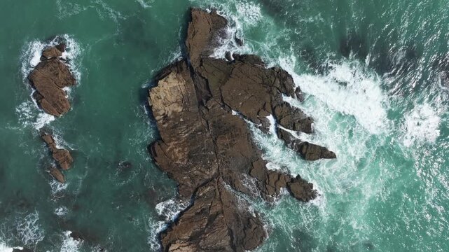 Aerial view of waves crashing against rugged rock formations in the ocean near the coast of Tunnel Beach, Waikato, New Zealand. The powerful sea meets the ancient stone.