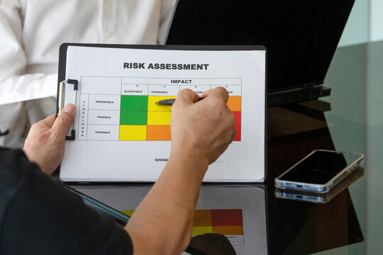 Over the shoulder shot of businessman holding pen over risk assessment report with colored matrix grid during office meeting.