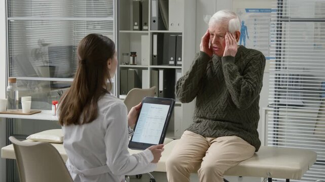 Medium full shot of elderly man complaining about headache to young female doctor with tablet during appointment in primary care office