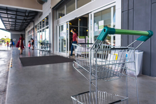 Empty metal shopping cart with green handle in front of supermarket. Shopping cart at entrance of department store. Chrome shopping cart in blurred mall background. Business and consumerism concept.
