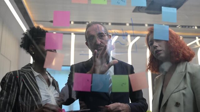 Multicultural business team brainstorming with sticky notes on glass wall, coworkers analyzing project strategy, creative coworkers collaborating in modern office, agile startup workflow concept.