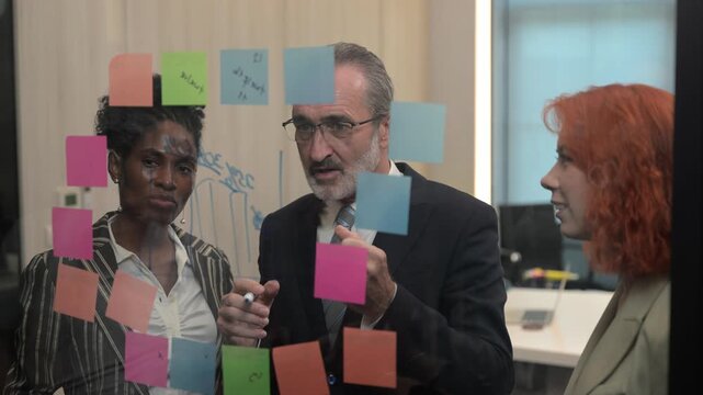 Multicultural business team brainstorming with sticky notes on glass wall, coworkers analyzing project strategy, creative coworkers collaborating in modern office, agile startup workflow concept.