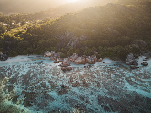 Aerial view of Anse Source d'Argent beach featuring iconic granite boulders, turquoise coral reefs, and lush tropical forest during golden hour in L'Union Estate, Seychelles.