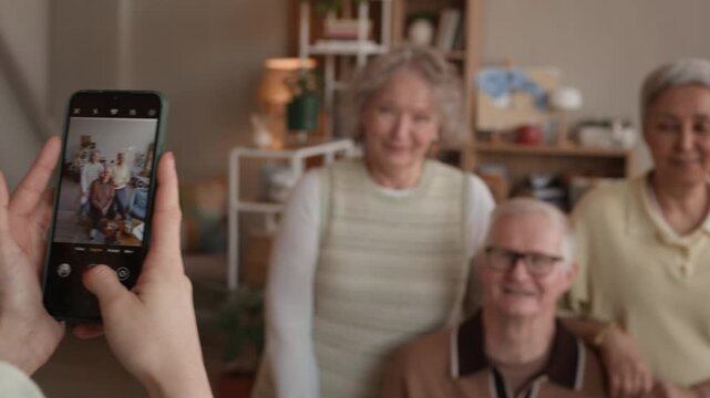Handheld shot of female caregiver taking photos of three senior friends on smartphone while working in nursing home