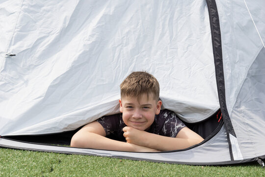 a smiling teenage boy peeks out from a gray camping tent while lying on his stomach and looking away with a joyful expression during a sunny day outdoors at a campsite or backyard