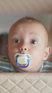 A baby is looking curiously over a crib with wide eyes. The baby holds a pacifier in their mouth and appears attentive to the surroundings.