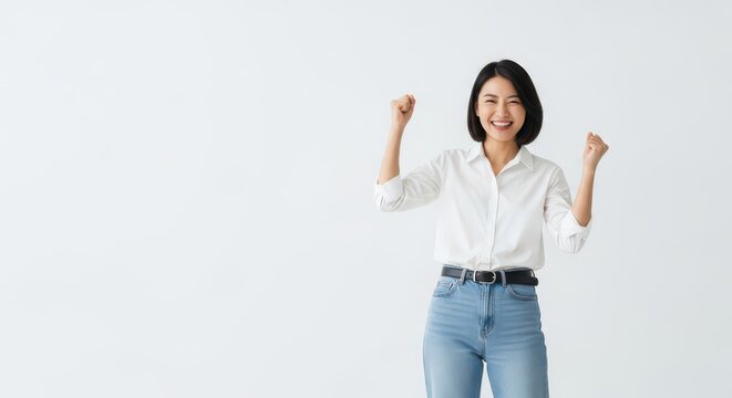 Excited young Asian woman celebrates success by raising her fists in victory against a plain background.