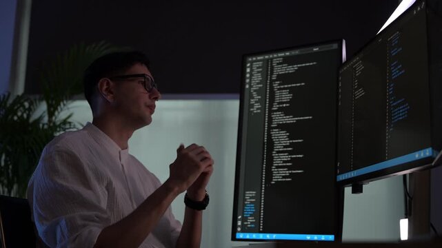 Focused young female software developer programming code on multiple monitors in a dark home office at night. Professional woman engineer working on cyber security and data science tech project.