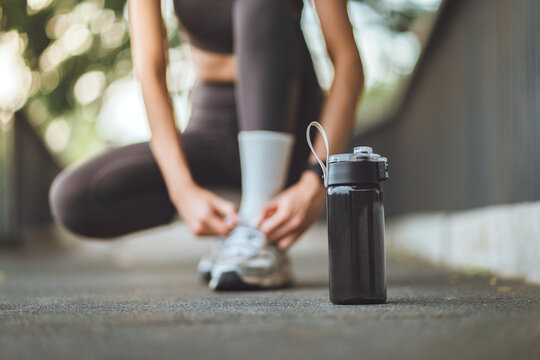 Asian young woman tying shoelaces in a park with a black water bottle in focus. Healthy lifestyle and fitness preparation concept for morning exercise, Wellness and sports marketing.