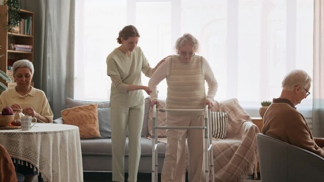 Long stab shot of young Caucasian caregiver smiling while assisting cheerful elderly woman with walkers, other nursing home residents resting in room