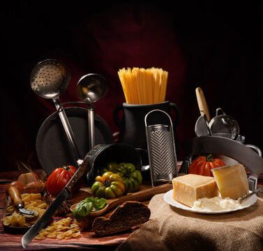 Still life, natura morta con pomodori su tavola imbandita con scenografia di vasi, pentole, utensili, calice di vino e brocca