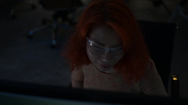 Focused female software developer coding at night in a modern workspace, wearing glasses and analyzing code on computer screen. Concept of programming, technology, cybersecurity, and digital innovatio