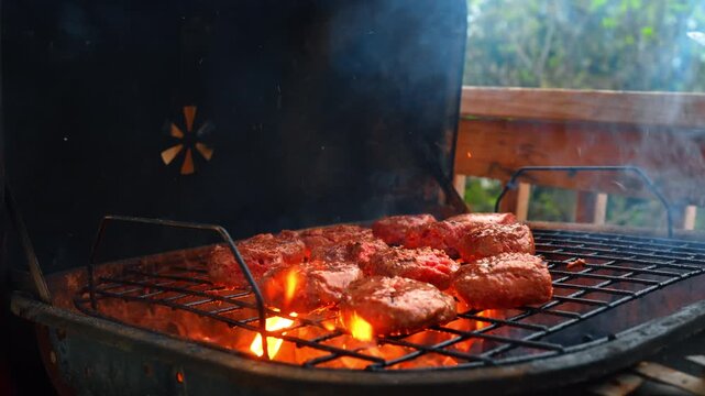 Beef burgers grilling over open flames on barbecue grill. Multiple meat patties cooking on a metal rack with bright orange fire in a black grill.