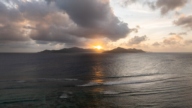 Aerial view of the golden sunset over the Indian Ocean with silhouettes of islands under a dramatic cloudy sky in Anse Reunion, La Digue, Seychelles.