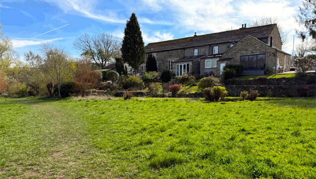 A rustic stone house sits comfortably in a lush green field under a bright blue sky. The surrounding trees and shrubs add a touch of nature's tranquility in Addingham, Ilkley, UK