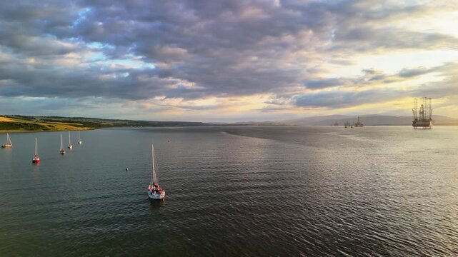 Aerial drone view of a lonely sailboat at anchor with offshore oil rigs at sunset, Cromarty Firth, Scotland