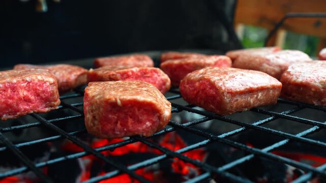 Raw meat patties on outdoor grill with glowing coals. Low angle side view of minced meat patties grilling on metal rack over hot burning charcoal in garden setting.