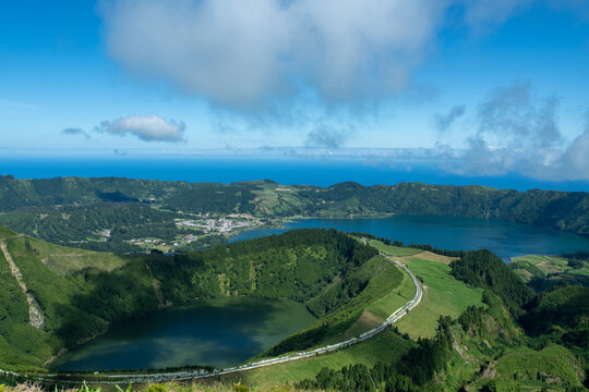Aerial panoramic view of volcanic twin lakes, green caldera slopes and coastal horizon on Sao Miguel Island, highlighting rural roads and lush landscapes. Sete Cidades, Ponta Delgada, Portugal.